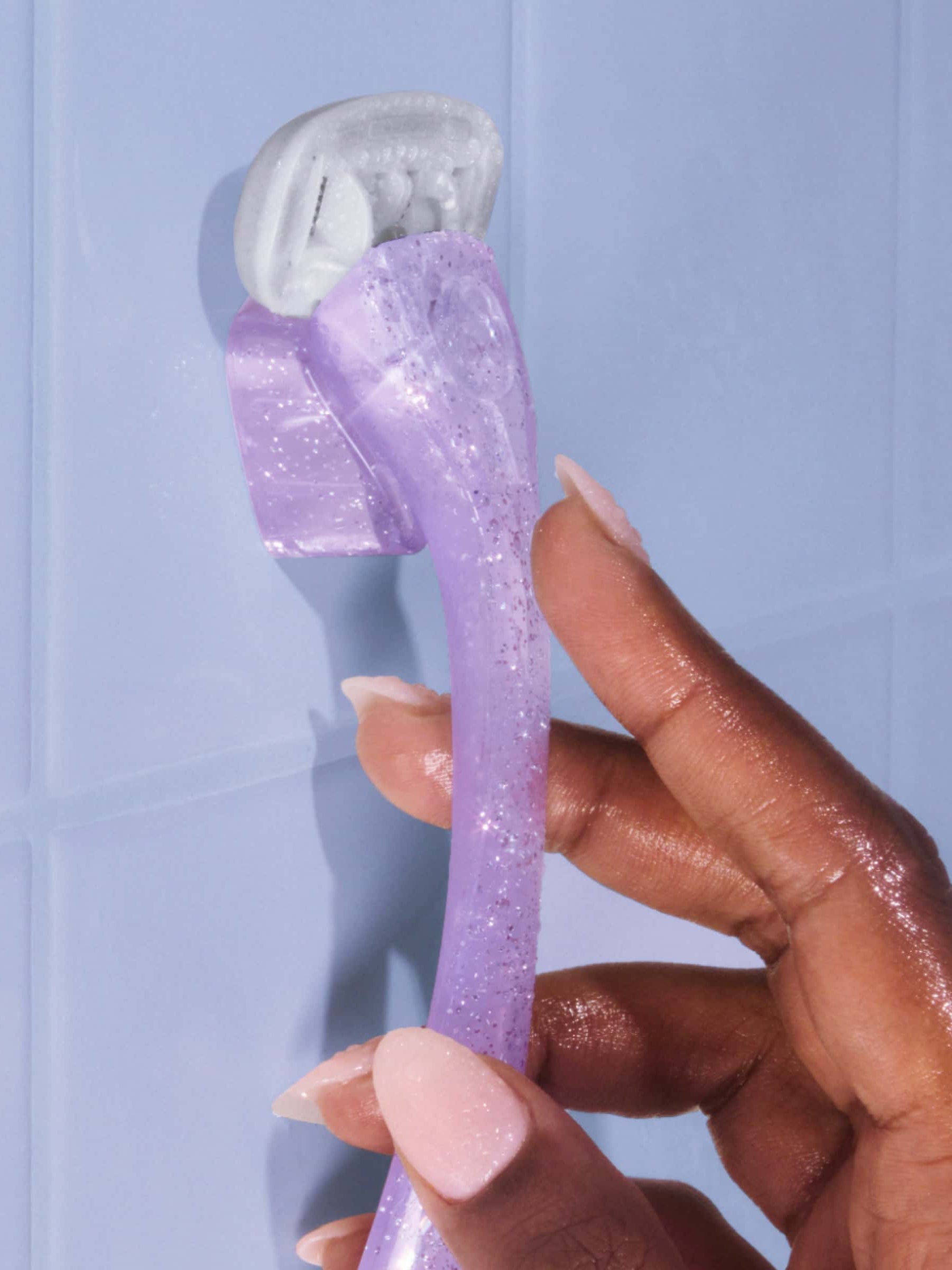 A hand holds a purple, sparkly razor with a white blade, positioned against a light blue tiled wall.