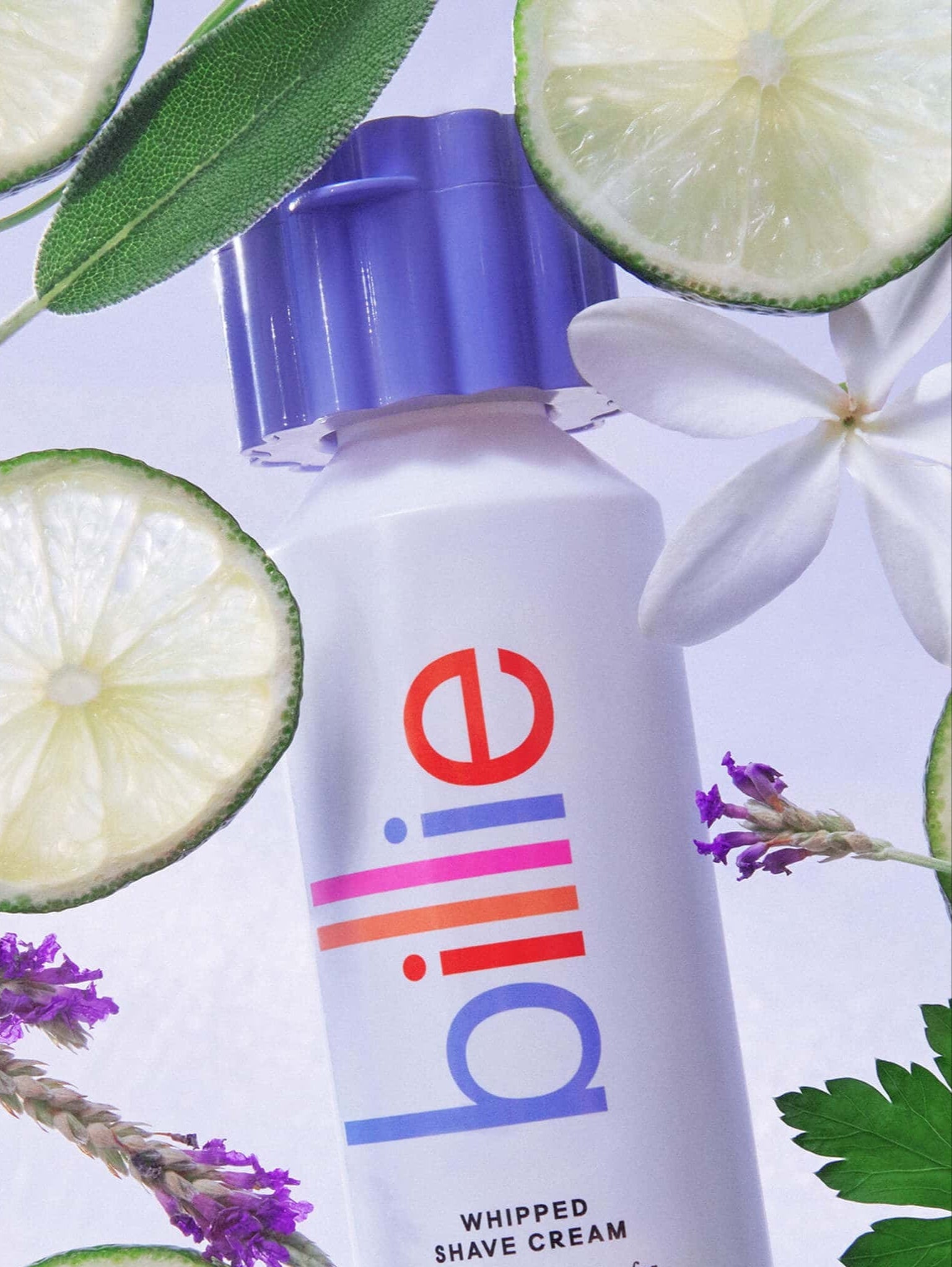 A white "billie" bottle with bold purple cap and text: "WHIPPED SHAVE CREAM," surrounded by limes, flowers, and leaves.