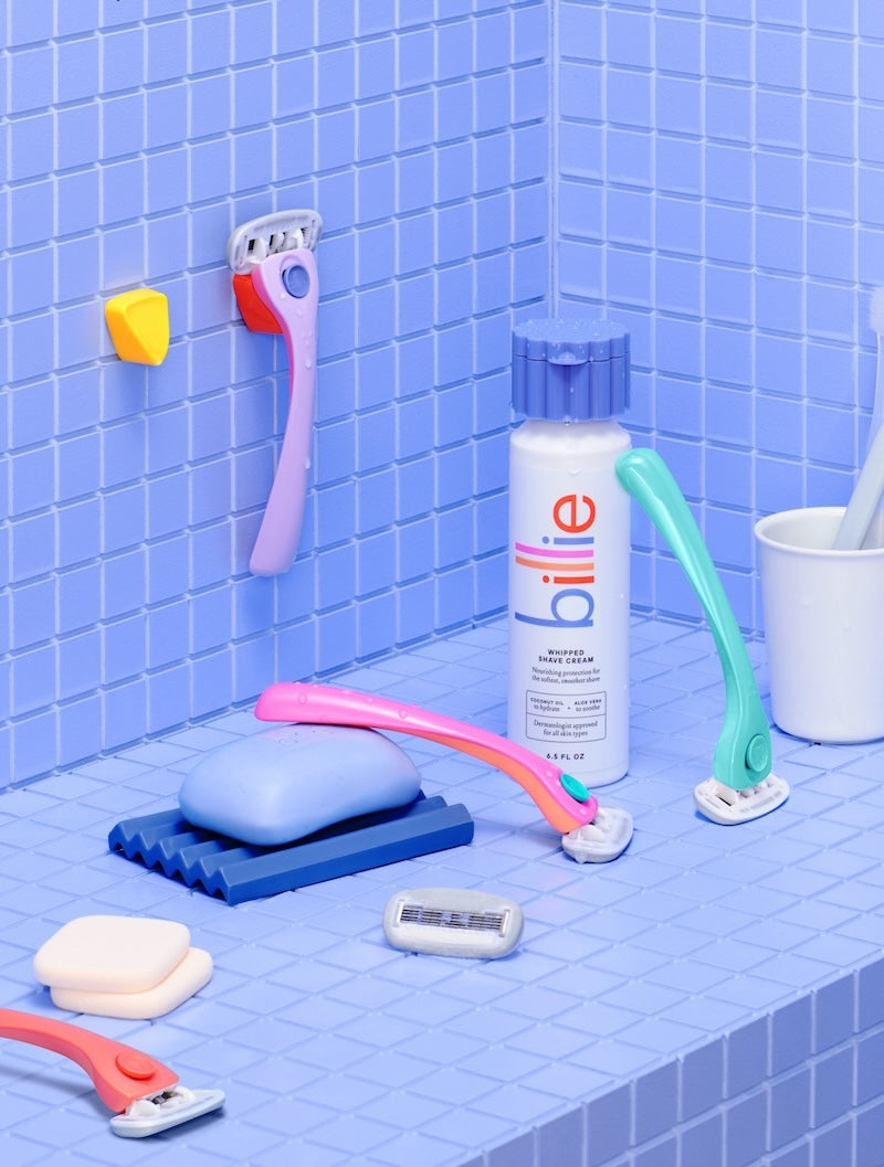 Toiletries on blue tiles: a purple razor hangs on the wall, near a blue "billie WHIPPED SHAVE CREAM" can and soap.
