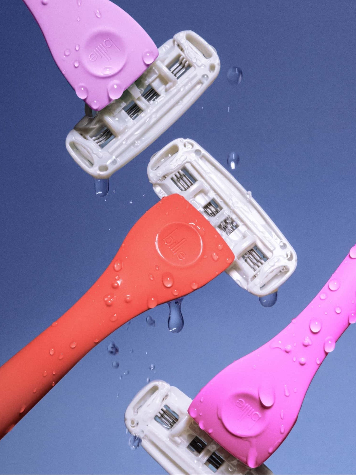 Four colourful "billie" razor heads intersect on a blue background, glistening with water droplets.