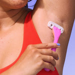 A person in a red top uses a pink razor to shave their underarm against a blue backdrop.