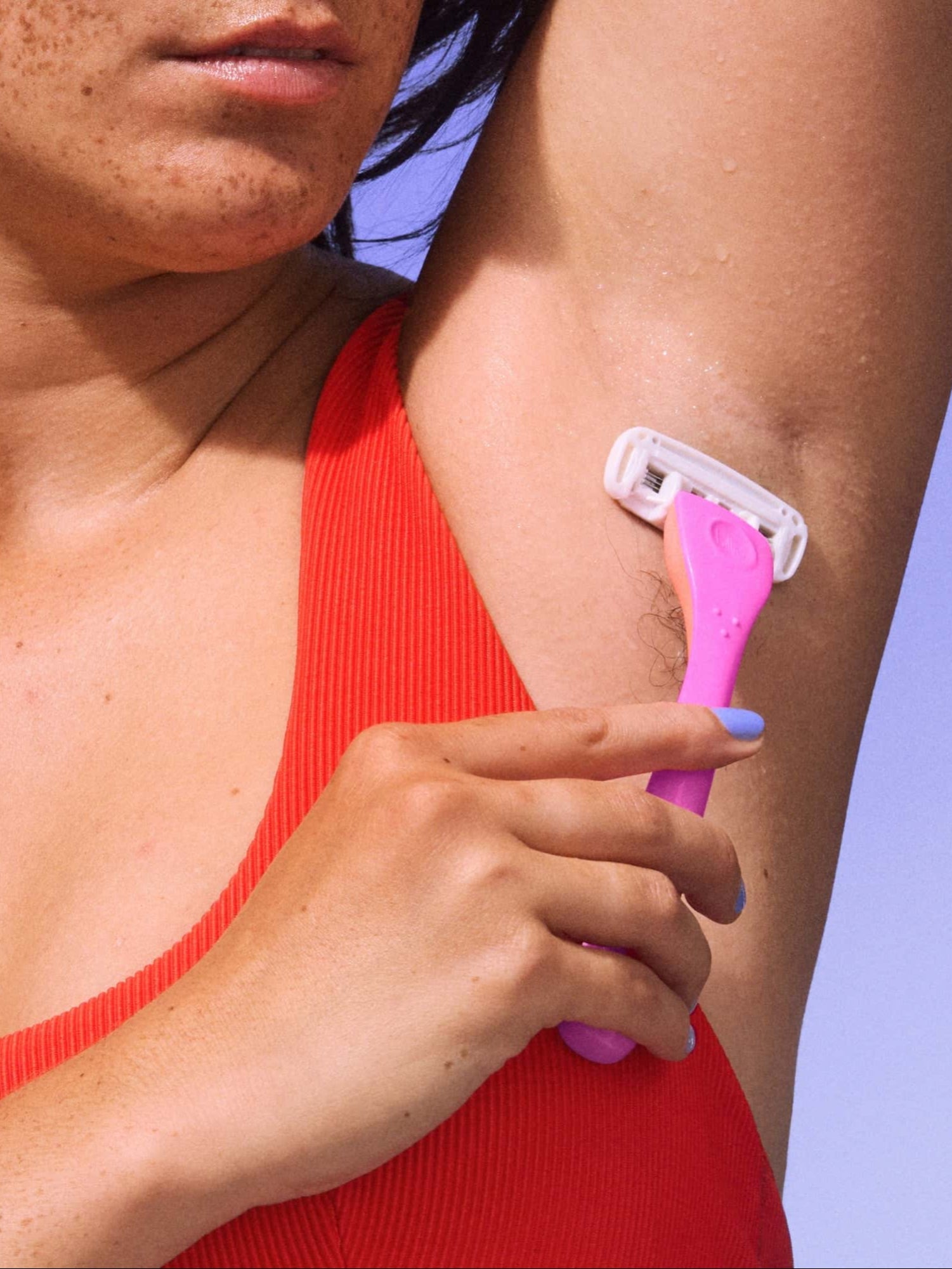 A person in a red top uses a pink razor to shave their underarm against a blue backdrop.