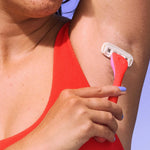 Hand holds pink razor, gently shaving an armpit; person wears red sleeveless top on light blue background.