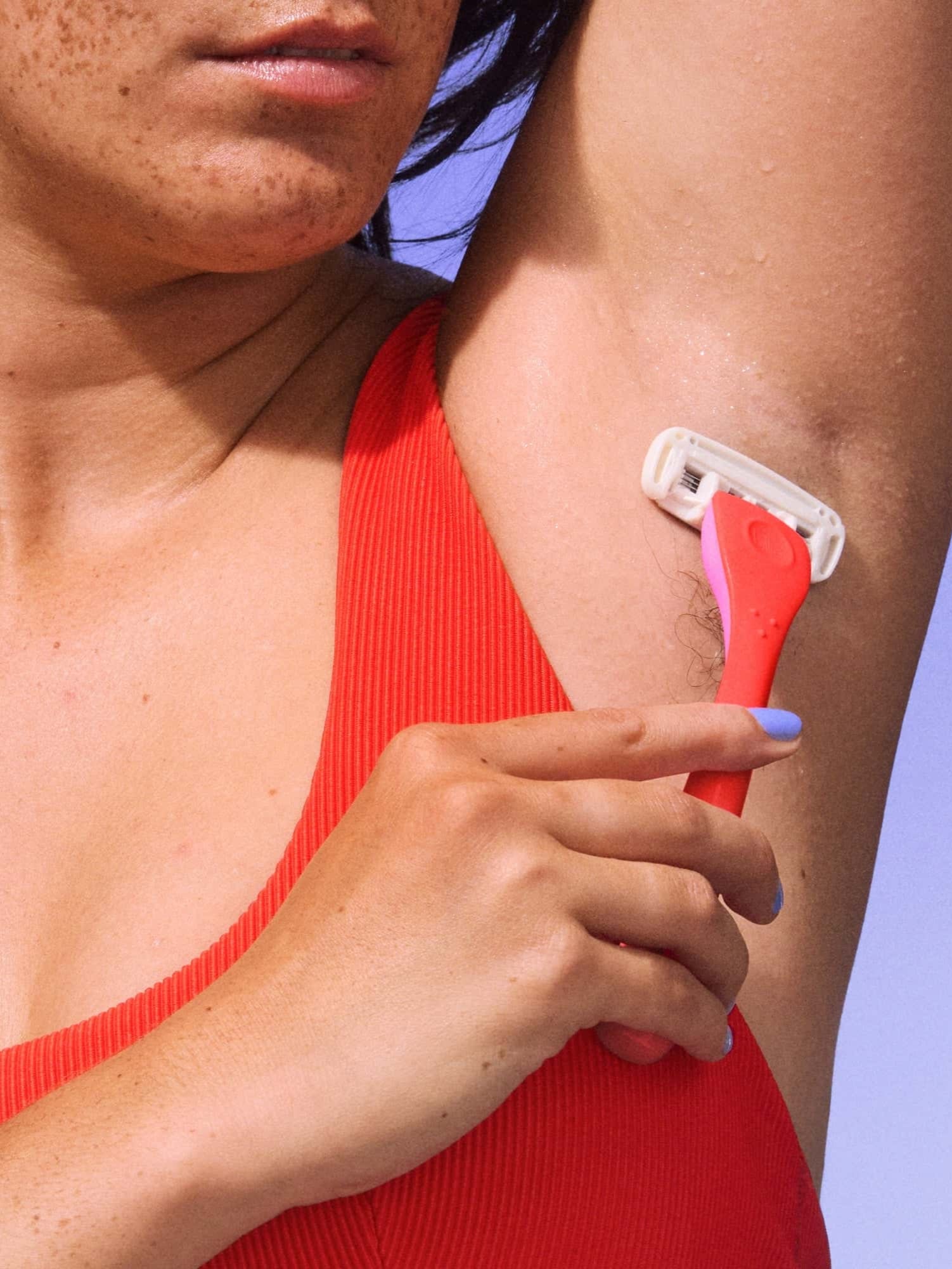 Hand holds pink razor, gently shaving an armpit; person wears red sleeveless top on light blue background.