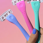 Hand with painted nails holds three "billie" razors—blue, pink, and green—against a pink background.