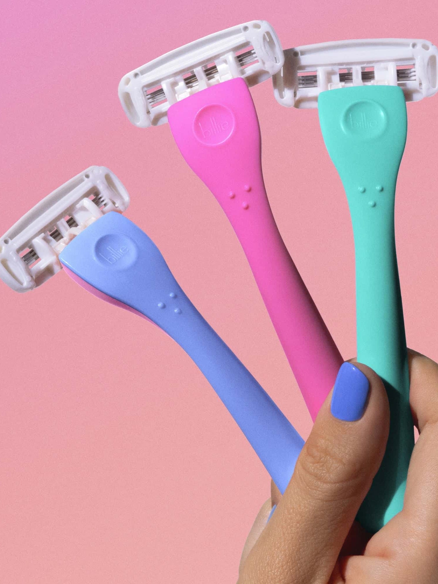 Hand with painted nails holds three "billie" razors—blue, pink, and green—against a pink background.