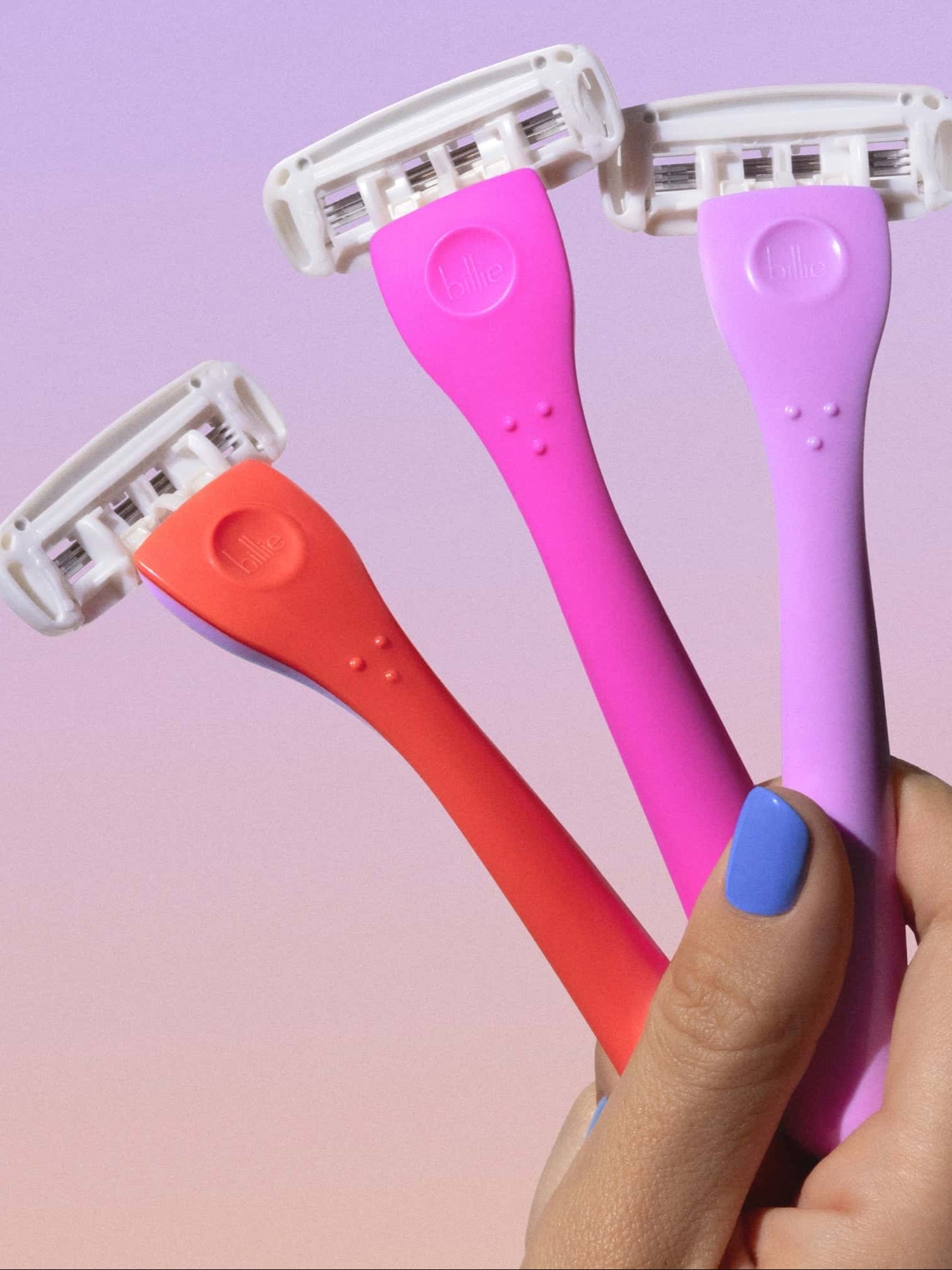 Three colorful razors in shades of red, pink, and purple are held in a hand with blue nail polish against a gradient background.