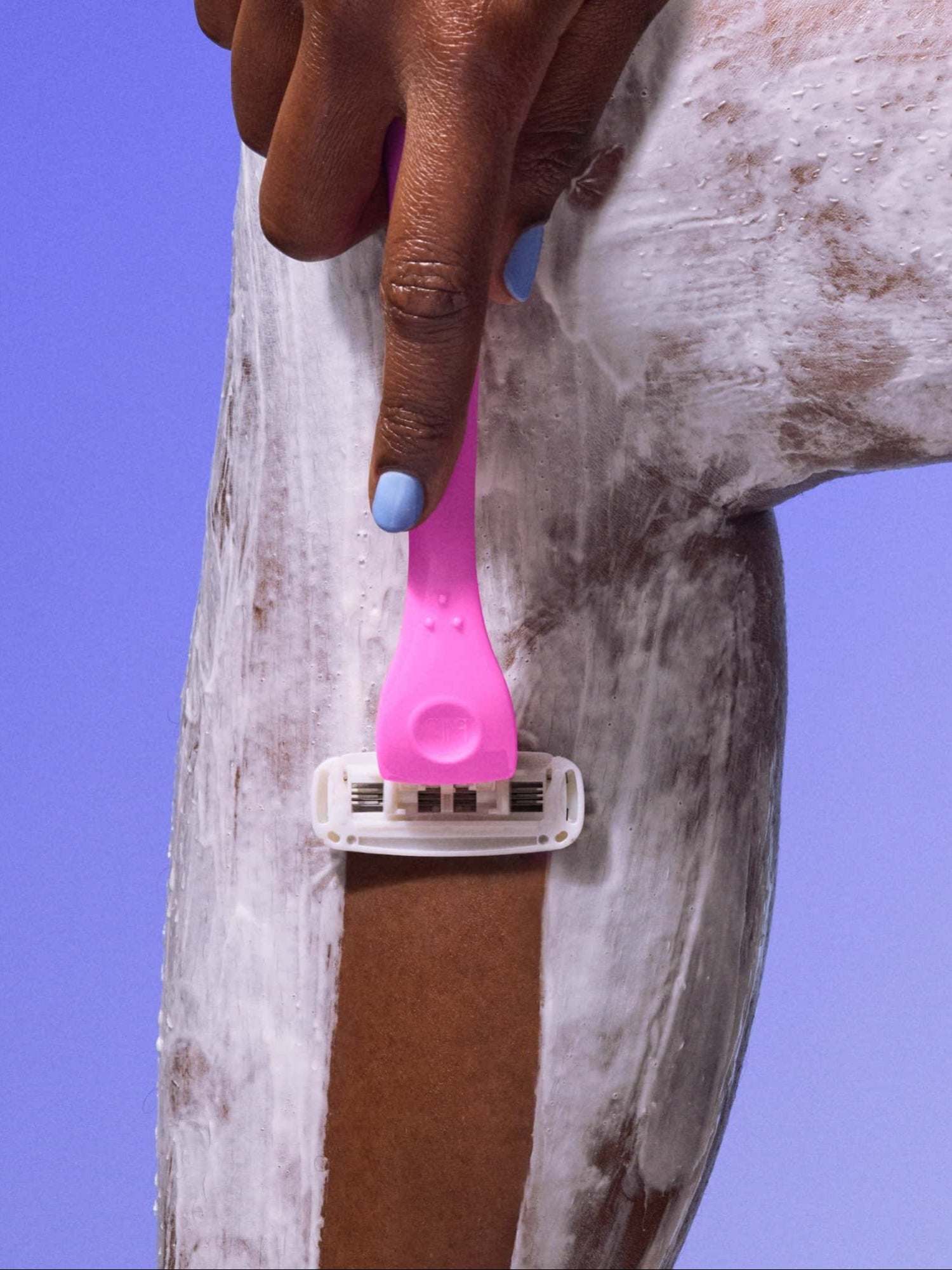 A pink razor shaves a person's soapy leg, removing shaving cream, against a blue-purple background.