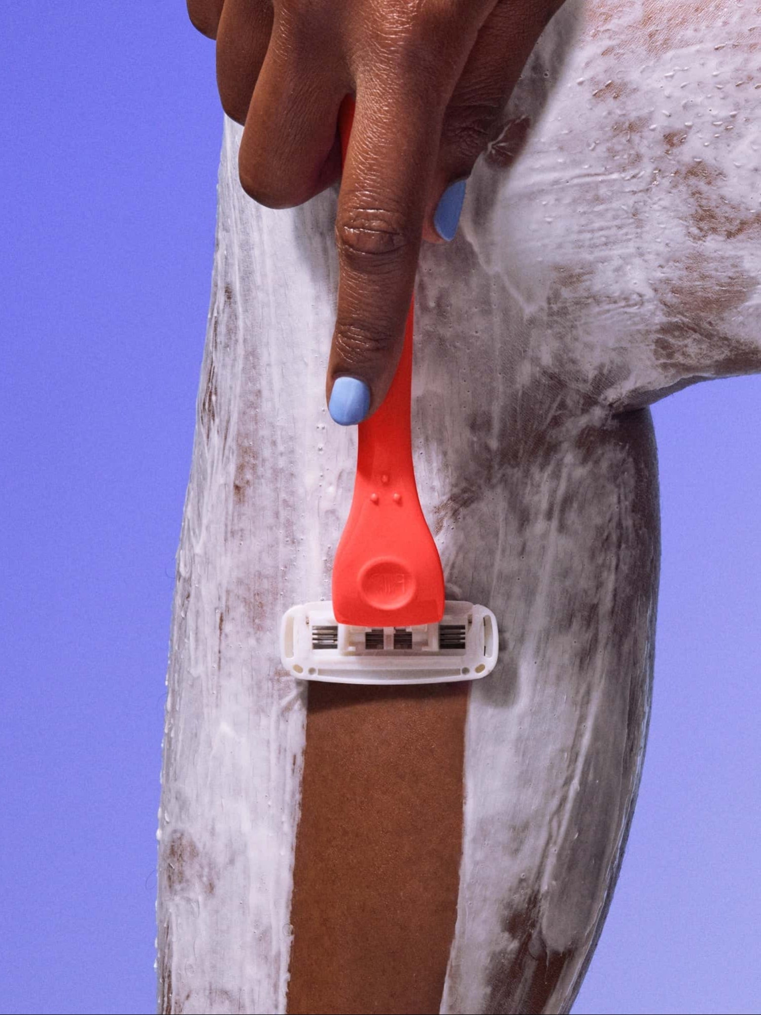A hand with blue nail polish uses a orange razor to shave a foam-covered leg against a purple background.