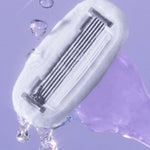 A razor head with multiple blades, water droplets glistening around it, set against a soft purple background.