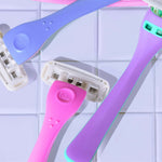 Three colorful razors with "billie" logos, in shades of blue, pink, and lavender, lie on a glossy, white tiled surface.