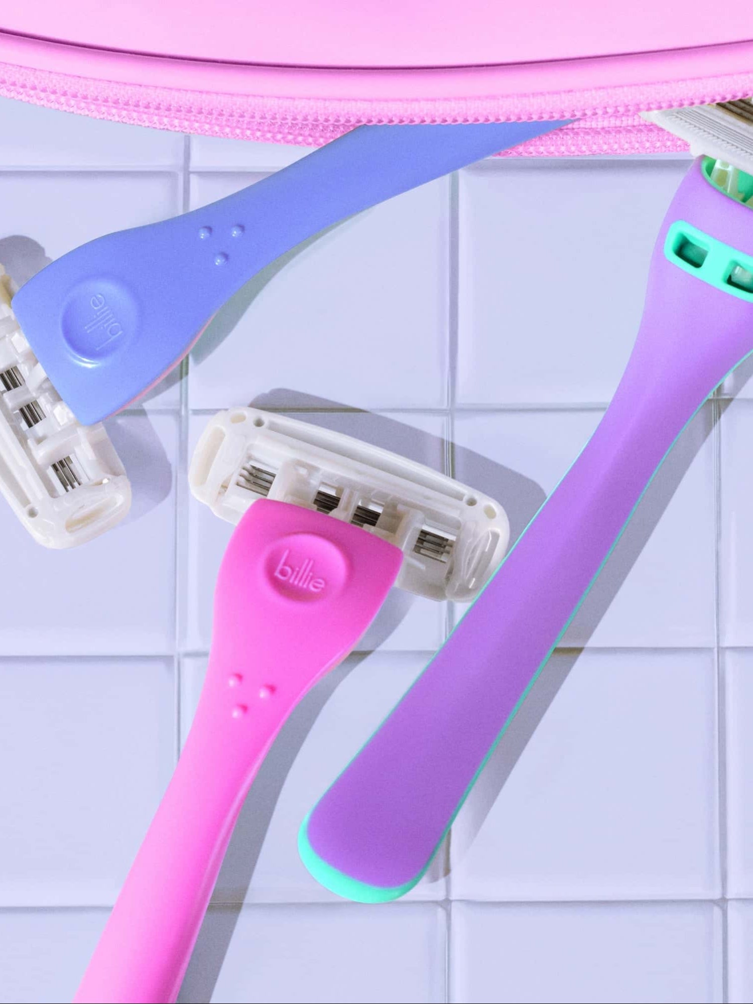 Three colorful razors with "billie" logos, in shades of blue, pink, and lavender, lie on a glossy, white tiled surface.