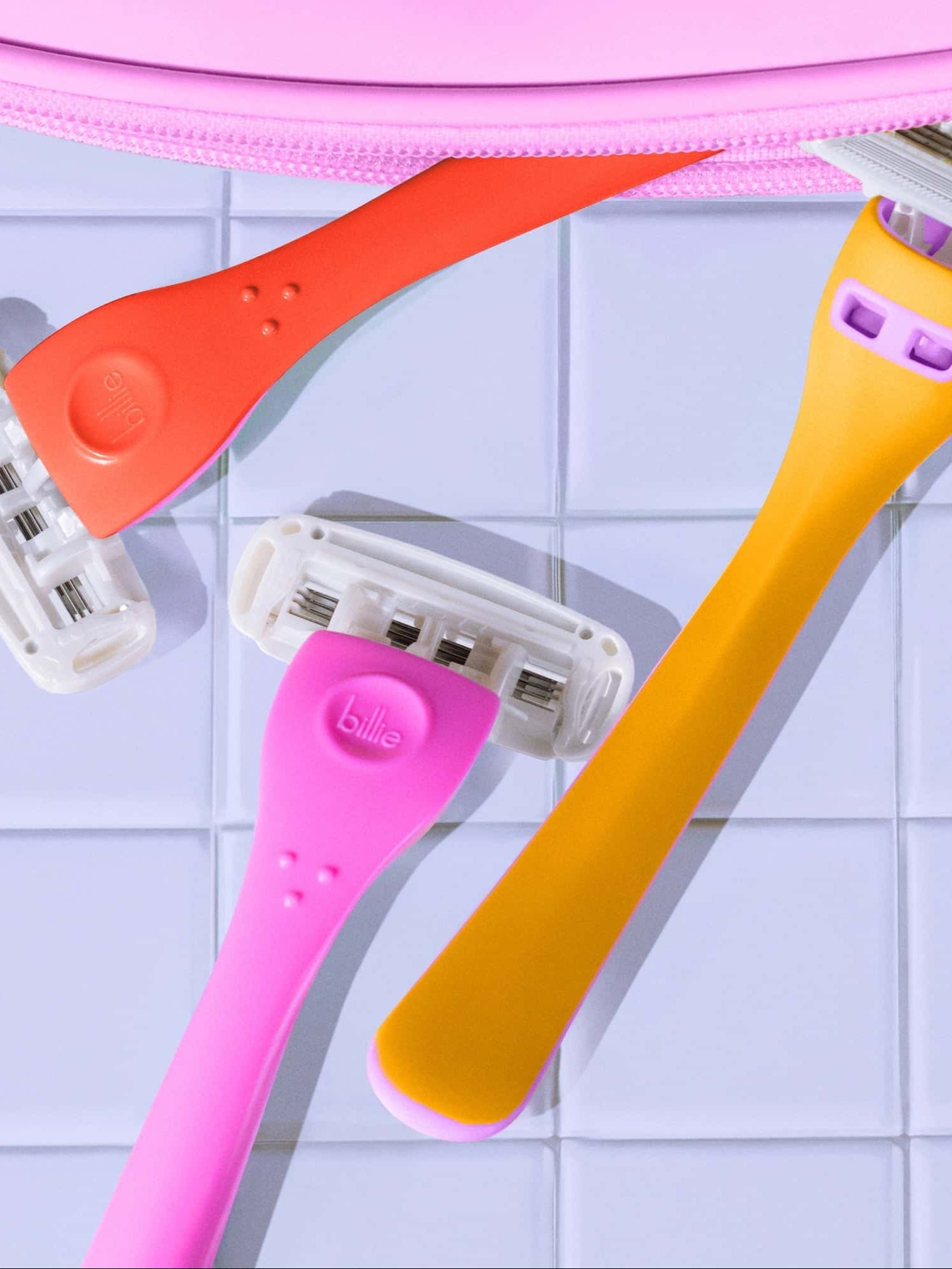 Three vibrant razors—orange, pink, and yellow—lie on a tiled surface near a pink zippered pouch. Text on the pink razor reads "billie.