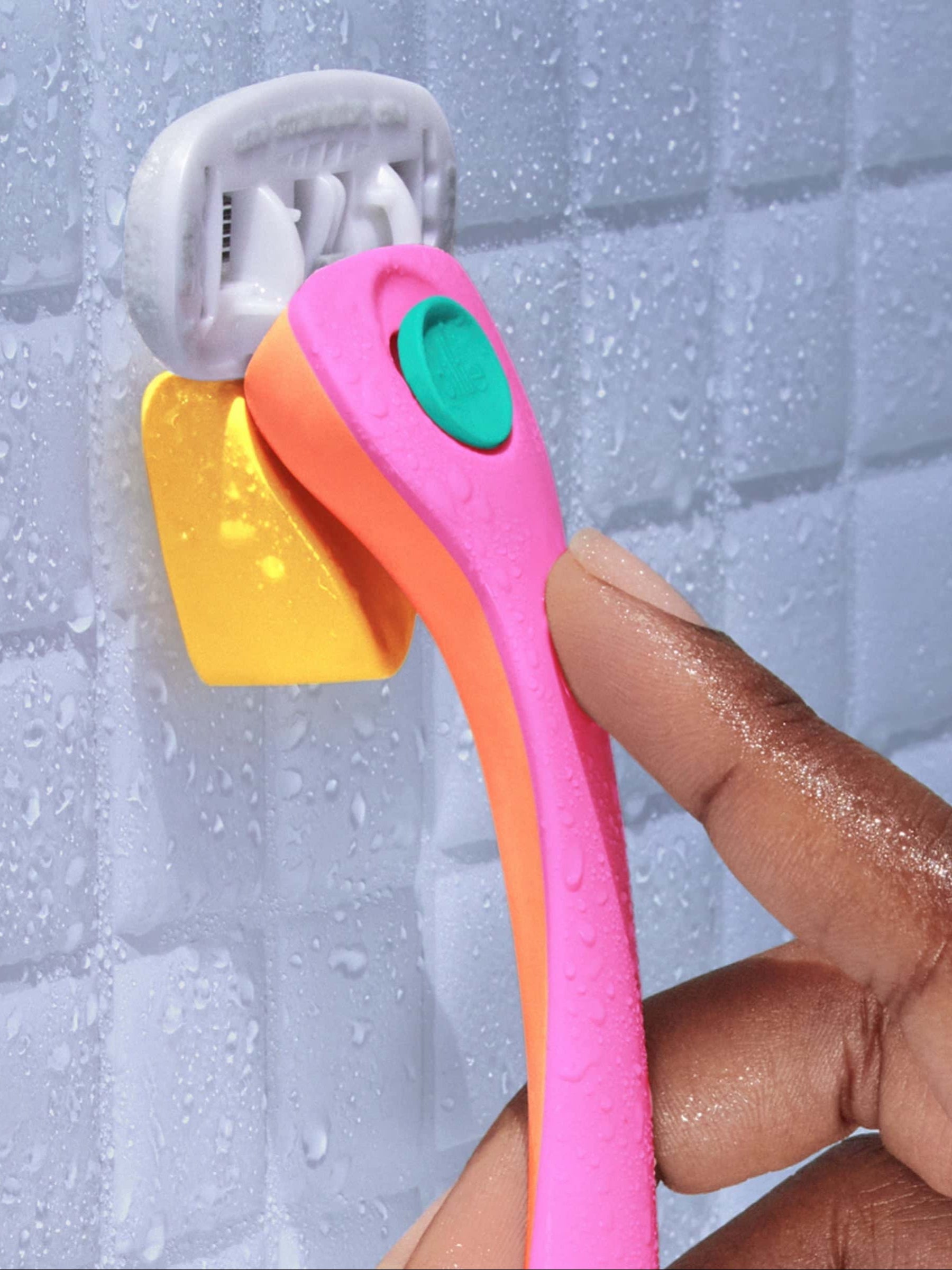 A colorful razor with a pink handle is being hung on a yellow hook, attached to a tiled bathroom wall wet with water droplets.