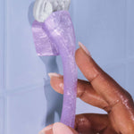 A hand holds a purple, sparkly razor with a white blade, positioned against a light blue tiled wall.