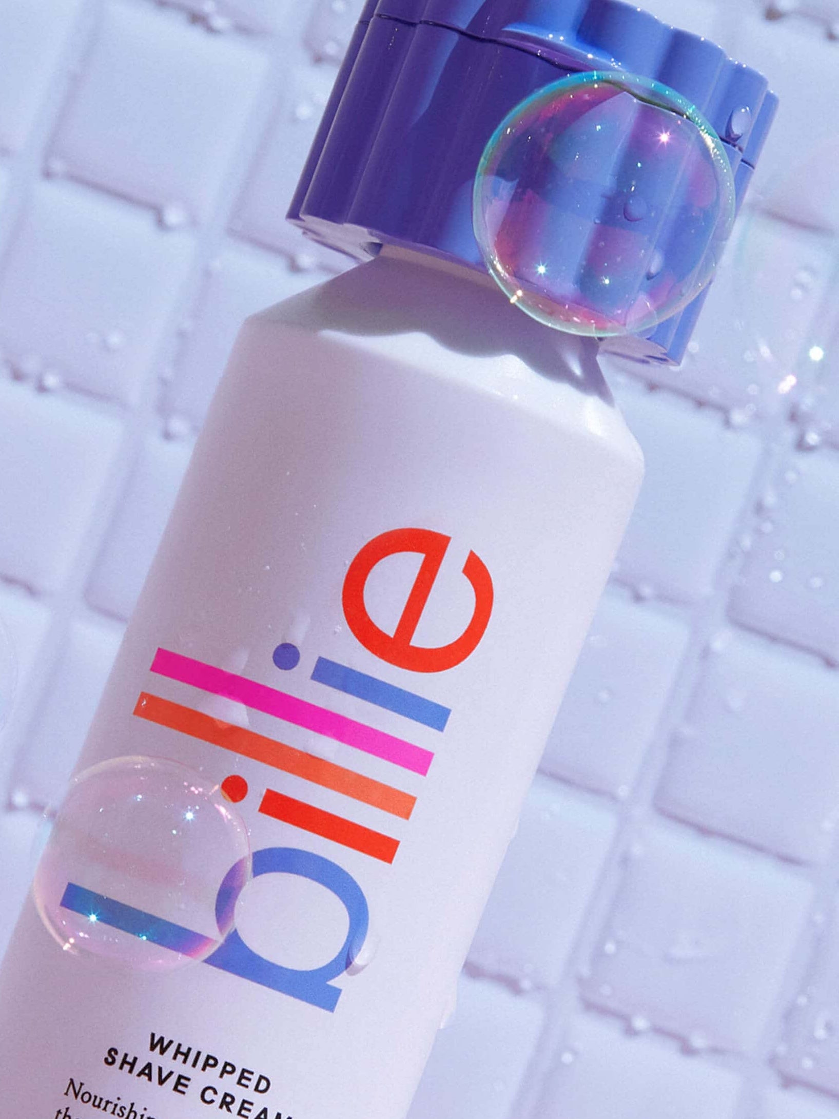 A "billie" shave cream can with blue cap, colourful branding, and soap bubbles on a tiled background.