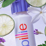 A white "billie" bottle with bold purple cap and text: "WHIPPED SHAVE CREAM," surrounded by limes, flowers, and leaves.