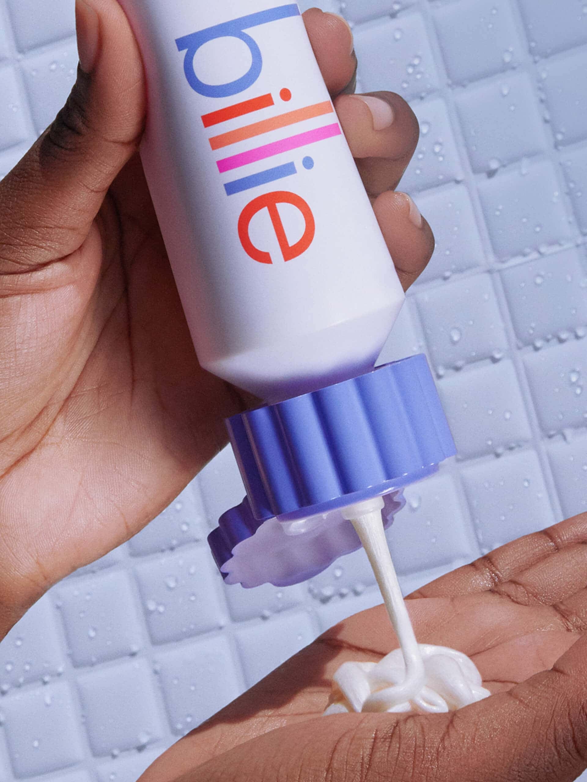 A hand dispenses shave cream from a white "billie" bottle onto another palm, against a wet tiled background.