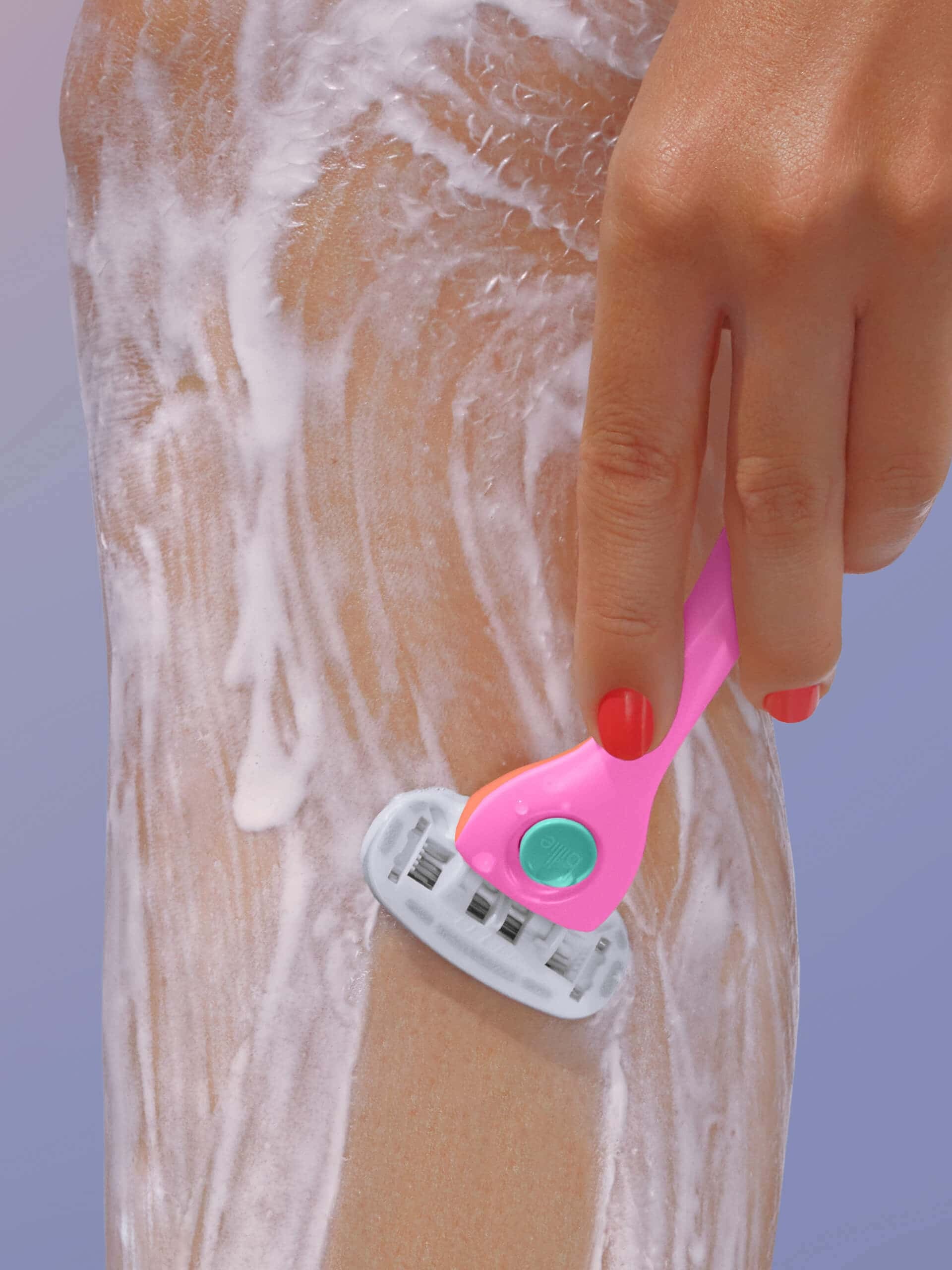 A hand with red nail polish uses a pink razor to shave a lathered leg against a soft blue background.