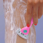 A hand with red nail polish uses a pink razor to shave a lathered leg against a soft blue background.