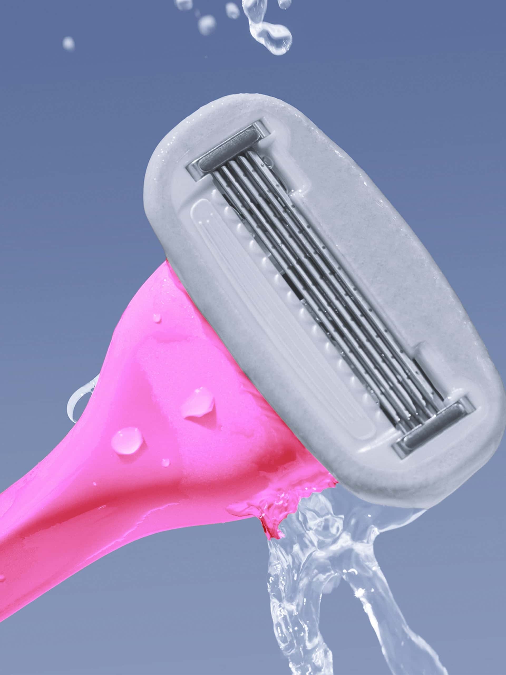 A pink multi-blade razor being rinsed, with water droplets on it and in the air, against a blue background.