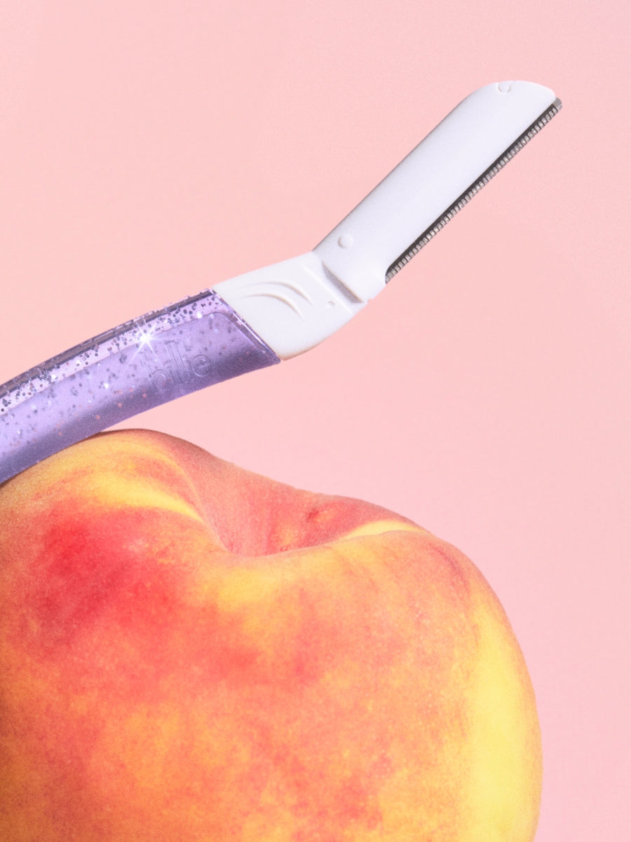 Purple glittery razor rests against a ripe peach, as if shaving it, on a soft pink background.