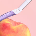 Purple glittery razor rests against a ripe peach, as if shaving it, on a soft pink background.
