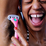 Smiling person shaves armpit with pink "billie" razor, wearing a striped top and showing joy.