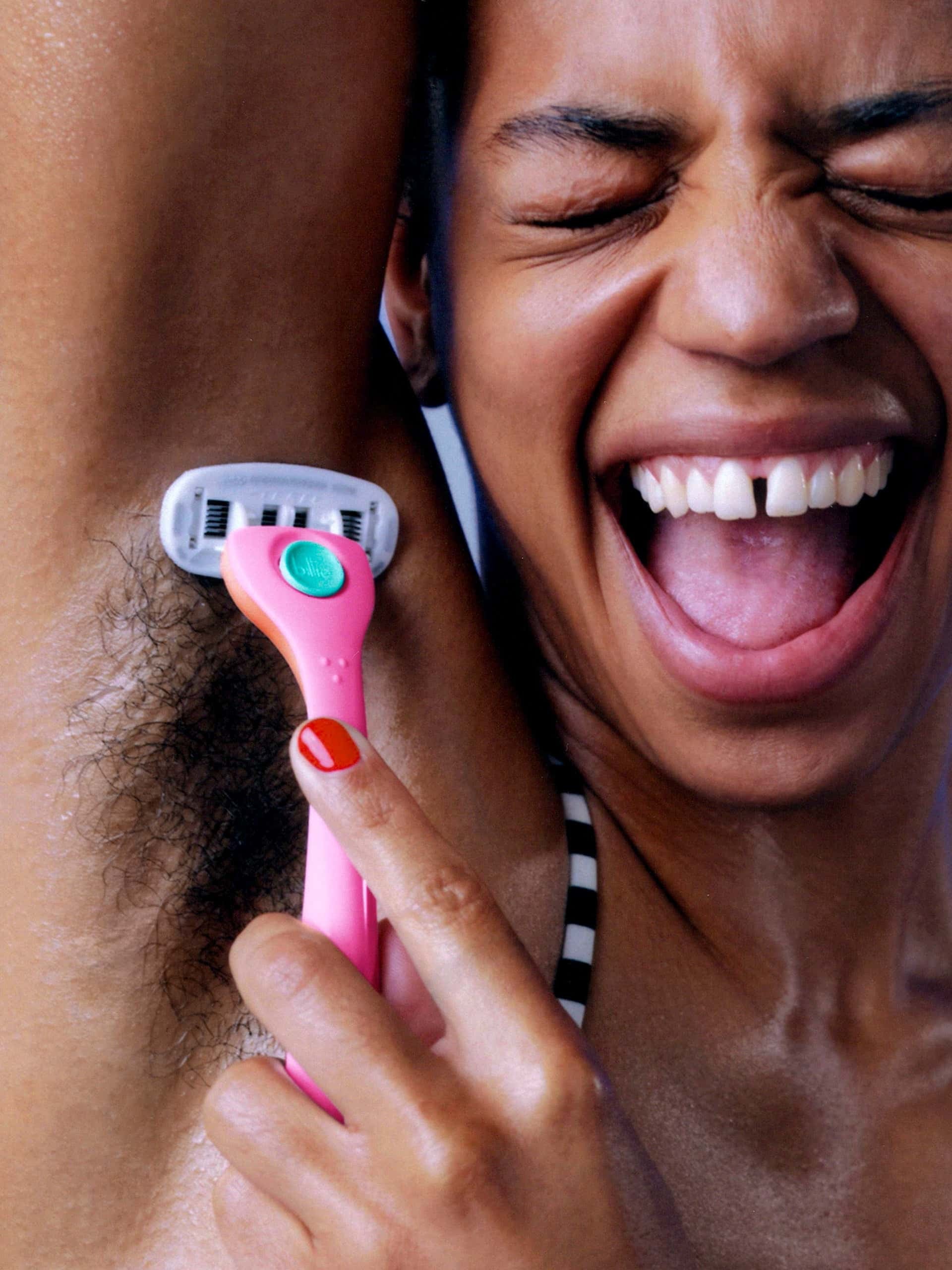 Smiling person shaves armpit with pink "billie" razor, wearing a striped top and showing joy.