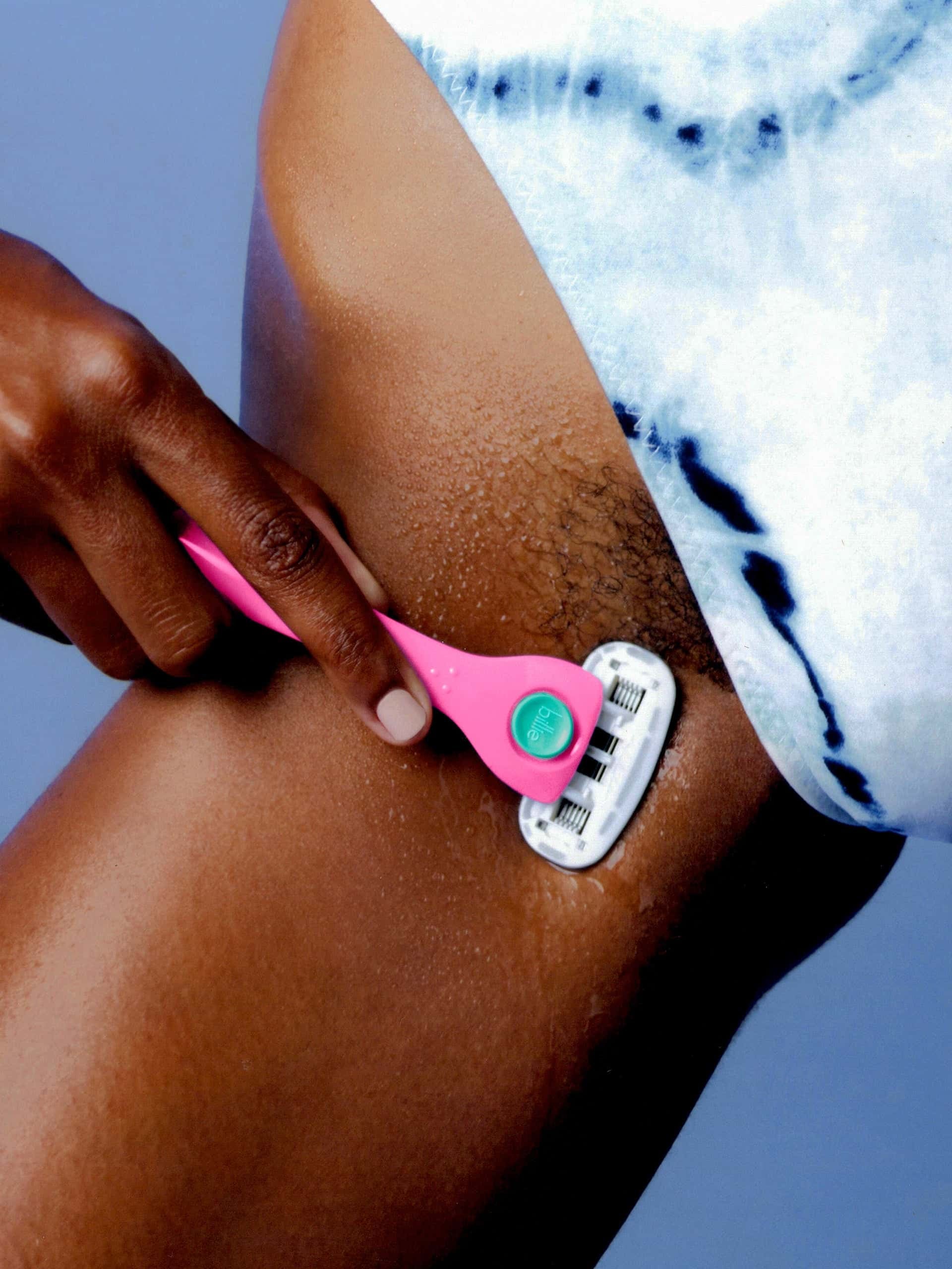A hand shaves a wet thigh with a pink "Billie" razor; blue tie-dye clothing is partially visible.