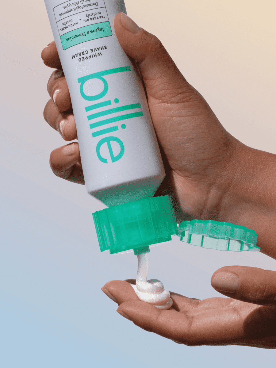 Hand dispenses "billie" Whipped Shave Cream from teal-capped tube into another, on soft gradient background.