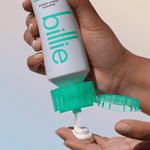 Hand dispenses "billie" Whipped Shave Cream from teal-capped tube into another, on soft gradient background.